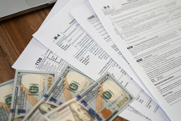 Tax forms and dollar bills spread out on a wooden desk near a laptop. Financial management, tax refund, and accounting concept. 