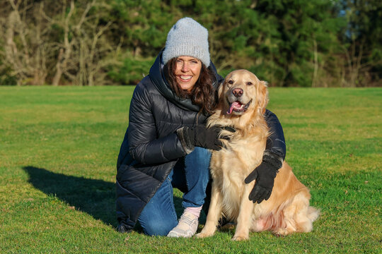 Adult woman wearing beanie cap and gloves for cold weather next to her friendly pet golden retriever dog outside on green grass. 