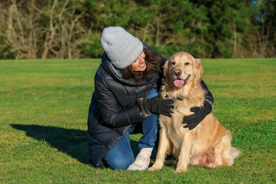 Adult woman wearing beanie cap and gloves for cold weather next to her friendly pet golden retriever dog outside on green grass. 
