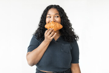 Portrait of happy black plus size female suffering eating disorder biting croissant secretively looking aside with cunning face, standing against white background, having serious food addiction