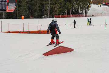 Naklejka premium Children skiers in snowpark in Dolomites Alps. Snow park box tricks. Skiers in park on a box. Winter jibbing in snwopark. Ski lessons on snowpark. Children learn to ski with an instructor.