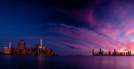 Fototapeta premium Panoramic view of Lower Manhattan and Jersey City during a vibrant sunset. The sky is painted with dramatic hues of purple, pink, and orange, reflecting softly on the calm waters of the Hudson River.