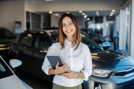 Portrait of a confident professional female car dealer standing in car showroom with tablet in hands.