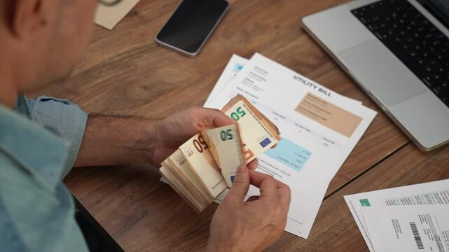 Mature businessman counts stack of euros reading utility bills at office table, male company accountant calculates fifty euro banknotes studying household expenses slow motion