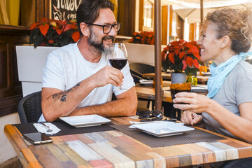 A man and a woman seated outdoors at a restaurant table, smiling at one another, holding a glass of wine and a soda, surrounded by a calm and relaxed atmosphere, while waiting for their ordered food
