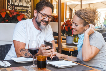 A couple of 50 years old with a beard and glasses for the man and blonde curly hair for the woman, enjoy a peaceful moment outdoors at a restaurant table while looking at funny content on their phone.