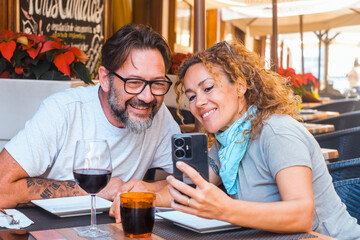 A couple of 48 years old enjoys a leisurely lunch outside at a restaurant, laughing together as the woman shares funny mobile content with her husband, in a peaceful outdoor setting under the sun.