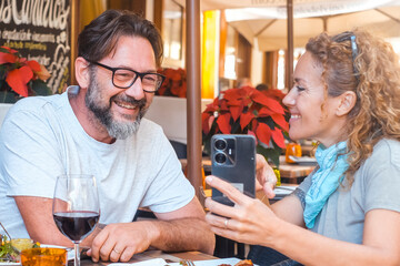 A couple of 48 years old enjoys a leisurely lunch outside at a restaurant, laughing together as the woman shares funny mobile content with her husband, in a peaceful outdoor setting under the sun.