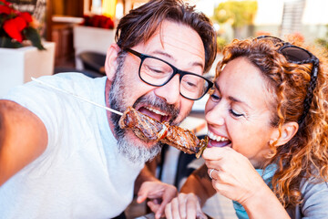 Two old friends seated at a restaurant table, enjoying a grilled meat skewer while smiling and laughing together. One of them is wearing glasses and the other has curly blonde hair, sharing a selfie.