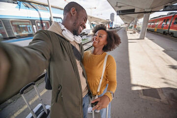 Joyful African American couple taking a selfie at a railway station before embarking on their train journey together. Travel happiness concept.