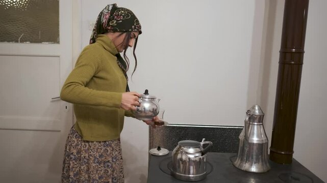 A young woman in a traditional village home pours tea from a kettle near a wood-burning stove.Wearing a headscarf, she reflects rural life, warmth, and hospitality, capturing simplicity and tradition.