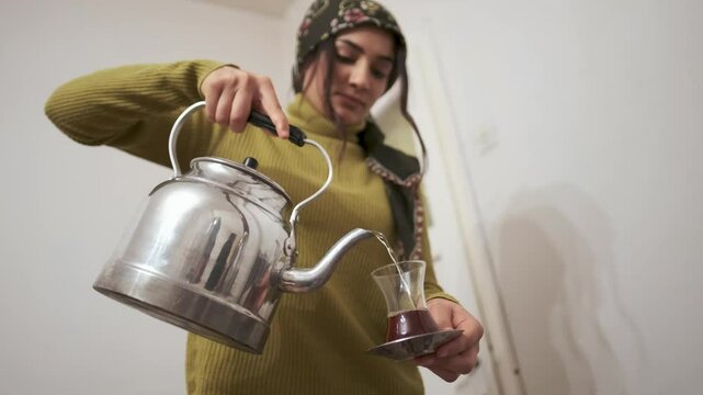 A young woman in a traditional village home pours tea from a kettle near a wood-burning stove.Wearing a headscarf, she reflects rural life, warmth, and hospitality, capturing simplicity and tradition.