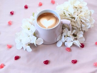 Coffee with Heart Latte Art in White Mug with Hydrangeas and Candy Hearts on Pink Background – Perfect for Valentine's Day Themes
