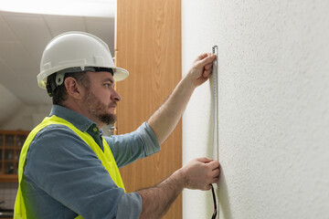 A construction worker in a white safety helmet and a bright yellow reflective vest measures with a...