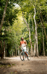 Obraz premium Viajero latino montando en bicicleta mirando a su alrededor, recorriendo la selva/jungla con su mochila, en México