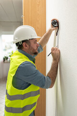 A construction worker in a white hard hat and a yellow reflective vest using a tape measure to take...