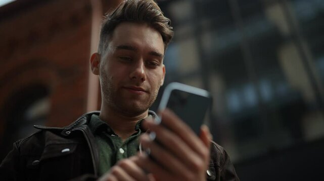 Close-up portrait of stylishly dressed young man standing on city street, with smile on face using smartphone to check messages, browse internet, stay connected enjoying urban environment, slowmo.