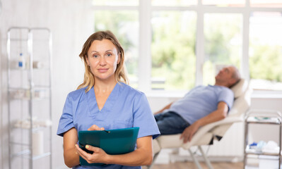 Positive middle-aged woman doctor posing in doctor's cabinet with documents and pen in hands