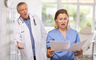 Upset shocked female medical worker in blue scrub standing in clinic office with papers in hands on...