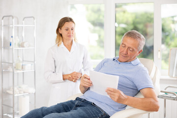 Obraz premium Elderly male patient signing informed consent form while lying on couch during medical consultation with female doctor. Compliance with legal requirements when receiving personalized medical care