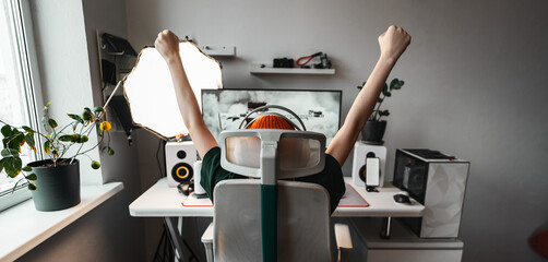 Young gamer girl in bright orange hat raises hands in excitement, celebrating victory in shooter game. Surrounded by professional gaming setup with monitor, headphones, gamepad. Joyful winner vibes