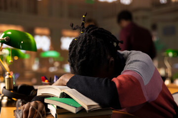 African american girl sleeping on books stack after a long night of studying for exams, struggling to finish college and pass university periodicals. Exhausted drowsy student napping due to overload.