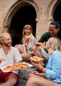 Fototapeta Vertical. Funny group of young people and they have fun eating pizza in the touristic city. Italian cheerful friends having food outdoors, laughing in a casual street setting. Vibrant urban background