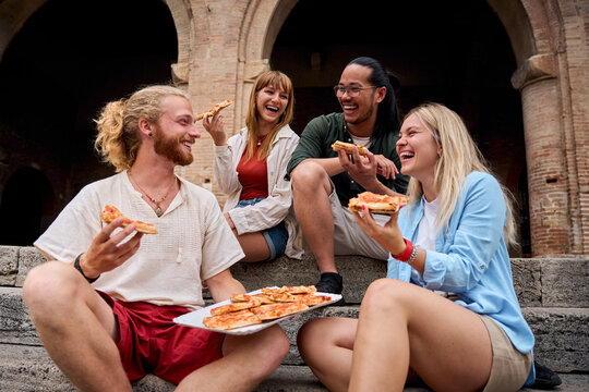 Fototapeta Funny group of young people and they have fun eating pizza in the touristic city. Italian friends having food outdoors, laughing in a casual street setting. Vibrant urban background and cheerful mood