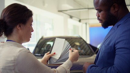 Fototapeta premium Saleswoman in dealership talking with client, offering color swatch samples choice, trying to grasp their preferences for vehicle. Agent providing customer with color wheel tinting selection, camera B