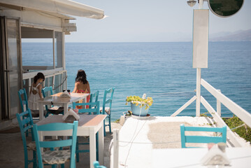 A young Asian woman wearing a colourful dress and her daughter walk towards a sunset viewpoint across chairs and tables overlooking the Aegean sea in a sunny summer day in  Crete, Greece