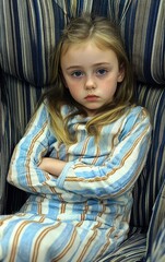 Fototapeta premium A young girl with long blonde hair sits in a comfortable striped chair, arms crossed and displaying a serious expression. Soft afternoon light fills the room, creating a calm atmosphere