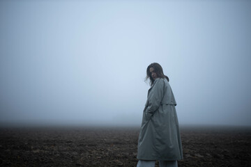 gloomy woman in coat walks alone in foggy field, existential crisis