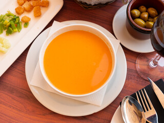 Cold soup gazpacho in bowl served with vegetables and baked breads on table in restaurant