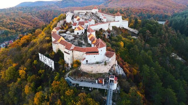 Drone footage of the iconic R&acirc;șnov Fortress perched atop a lush hill, showcasing its medieval architecture and stunning panoramic views of the surrounding Romanian countryside.