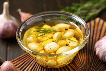 Glass bowl of fresh garlic oil with rosemary and peppercorns on wooden background, closeup