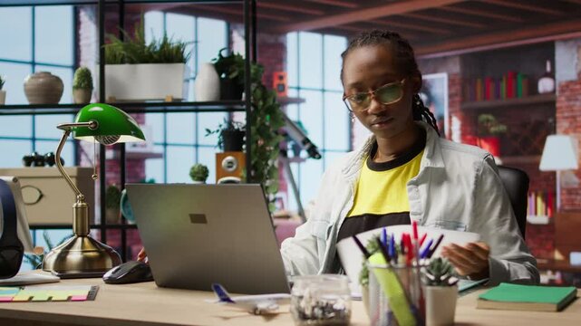 Teleworker using laptop in home office to crosscheck paper documents with online company files. Remote worker typing on notebook, inputting paperwork documentation data, camera A