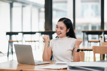 Fototapeta premium A joyful young woman celebrates success while working on her laptop in a bright, modern office setting.