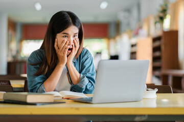 A young woman in a library looks surprised and excited while using a laptop, surrounded by books and natural light.