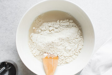 Overhead view of bread dough being mixed in a white bowl, top view of homemade bread dough being mixed on white granite countertop, process of making artisan bread