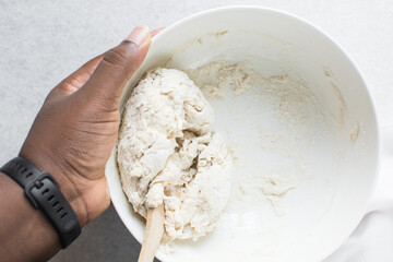 Overhead view of bread dough being mixed in a white bowl, top view of homemade bread dough being mixed on white granite countertop, process of making artisan bread