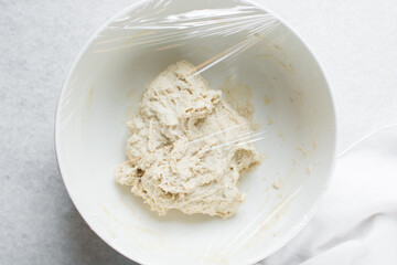 Overhead view of bread dough in a whiite mixing bowl covered with plastic wrap, dough rising in a ceramic bowl, process of making artisan bread