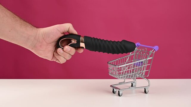 Man putting black sex toy in mini shopping cart on pink background. 