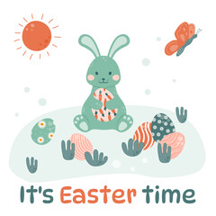 Easter bunny is sitting on the grass with colorful eggs and holding an Easter egg in his paws.