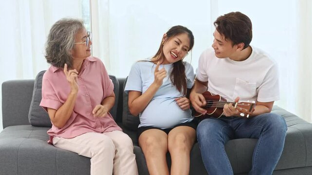 Asian family mother in law son in law and pregnant daughter close to birth happy family together son in law playing guitar singing sitting on sofa dancing happily in living room at home happy.