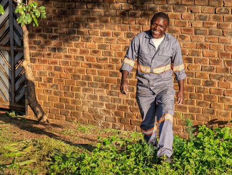 Smiling African man in reflective gray workwear, walking near a rustic brick wall in bright sunlight, outdoors with greenery, industrial and casual setting, South Africa - Powered by Adobe