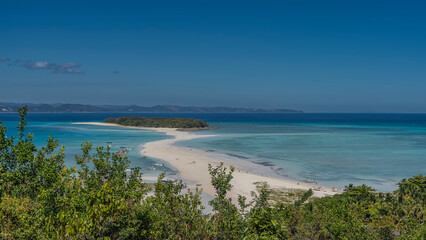 Beautiful tropical postcard landscape. A green island in the aquamarine ocean. Silhouettes of people on a winding sandbar. Boats near the shore. Green vegetation in the foreground. The blue sky. 