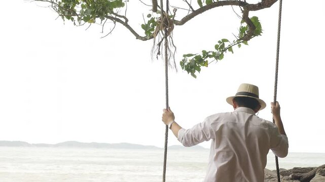 Man in white shirt and hat sitting on a tree swing by the beach, enjoying the serene view of the sea and horizon under a canopy of green leaves