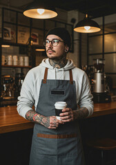 A tattooed barista in a cozy caf&eacute; wearing glasses, a beanie, and an apron, holding a takeaway coffee cup against a stylish coffee shop backdrop.