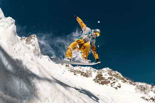 Professional male snowboarder jumps in powder snow from kicker against white snow capped mountains and blue sky. Freeride concept at ski resort