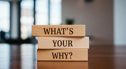 Three wooden blocks stacked vertically read "WHAT'S YOUR WHY?" in dark brown sans-serif font. Set on a dark surface with a blurred bright background,  for motivation, purpose, self-reflect